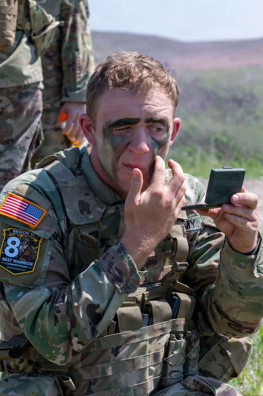A U.S. Army soldier in MultiCam (OCP) uniform is applying camouflage face paint while looking into a small compact mirror. He is wearing tactical gear and a patch identifying him as a participant in the Region VI Best Warrior Competition. The setting appears to be an outdoor training or competition area with hilly terrain in the background.