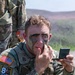 A U.S. Army soldier in MultiCam (OCP) uniform is applying camouflage face paint while looking into a small compact mirror. He is wearing tactical gear and a patch identifying him as a participant in the Region VI Best Warrior Competition. The setting appears to be an outdoor training or competition area with hilly terrain in the background.