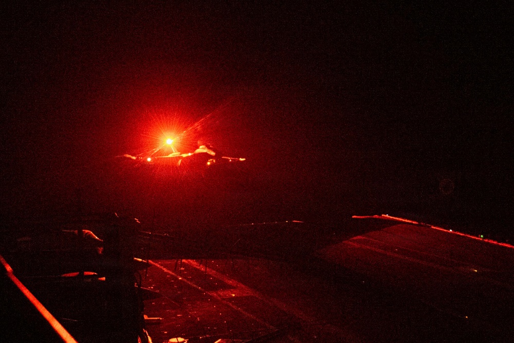 A nighttime image showing what appears to be a military jet aircraft launching or departing from what appears to be an aircraft carrier flight deck. The scene is dominated by intense red/orange lighting, likely from afterburner exhaust or deck lighting. The extremely low-light conditions and red color cast make fine details difficult to discern.