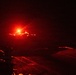 A nighttime image showing what appears to be a military jet aircraft launching or departing from what appears to be an aircraft carrier flight deck. The scene is dominated by intense red/orange lighting, likely from afterburner exhaust or deck lighting. The extremely low-light conditions and red color cast make fine details difficult to discern.