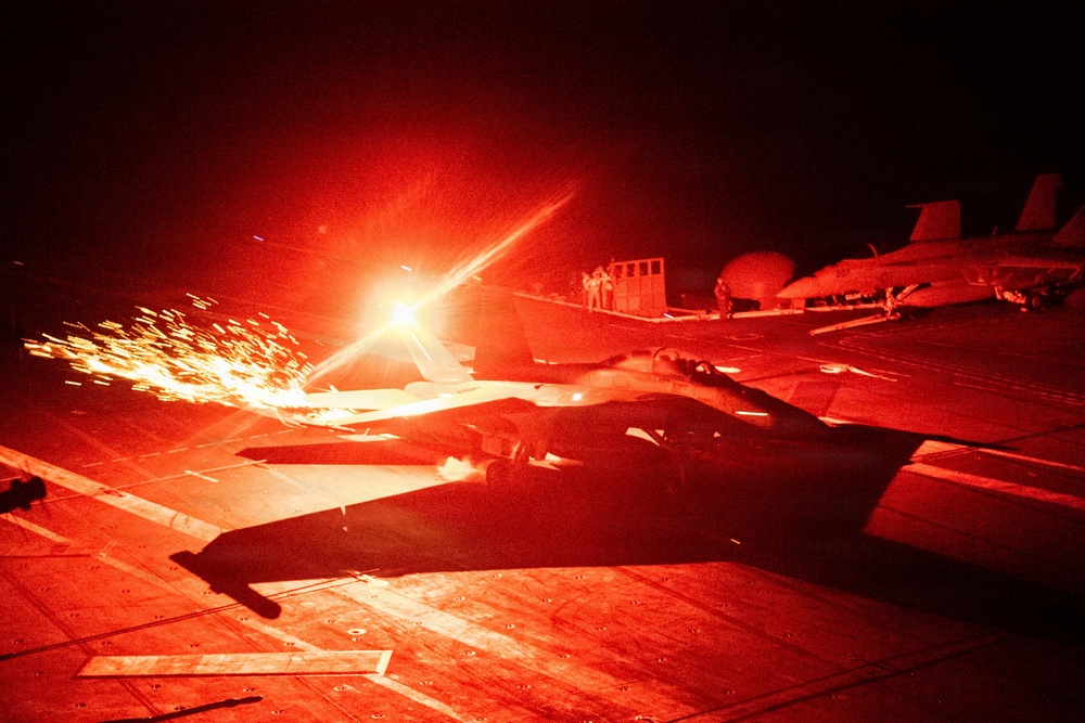 A nighttime scene on what appears to be an aircraft carrier flight deck, dramatically illuminated by intense red lighting and bright sparks or afterburner exhaust from a military jet aircraft. The image captures a jet launch or engine ignition event, with sparks and light streaks visible against the dark sky. Additional parked aircraft and deck personnel are visible in the background.