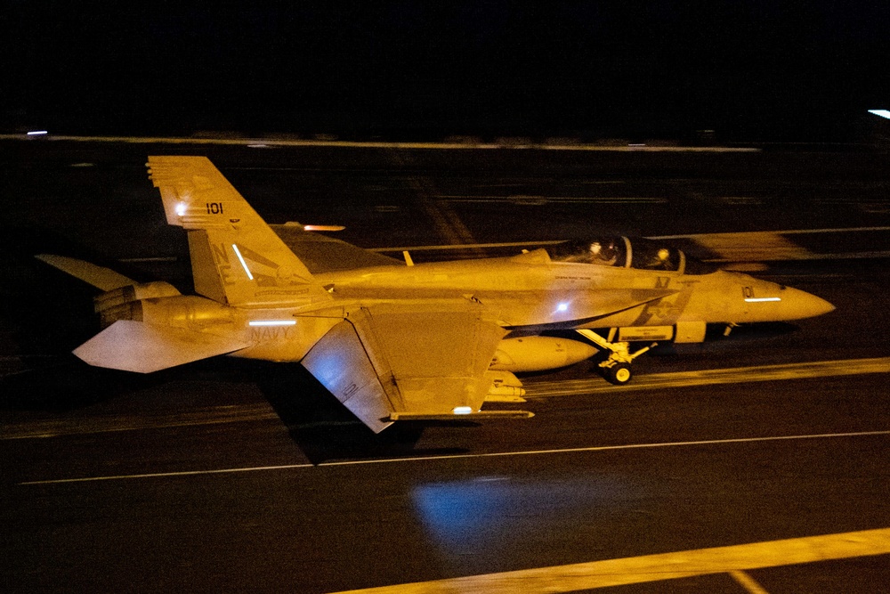 A U.S. Navy F/A-18 Super Hornet (modular variant) with tail number 101 is photographed at night on what appears to be an aircraft carrier flight deck. The aircraft is illuminated by artificial lighting against a dark background, showing 'NAVY' markings on the fuselage and an 'NJ' or similar tail code. The image captures the aircraft in a low-light operational environment, likely during night flight deck operations.