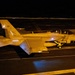 A U.S. Navy F/A-18 Super Hornet (modular variant) with tail number 101 is photographed at night on what appears to be an aircraft carrier flight deck. The aircraft is illuminated by artificial lighting against a dark background, showing 'NAVY' markings on the fuselage and an 'NJ' or similar tail code. The image captures the aircraft in a low-light operational environment, likely during night flight deck operations.