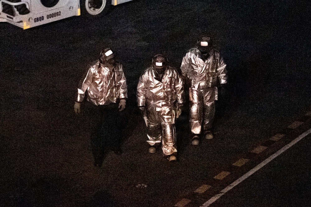 Three individuals wearing reflective silver/aluminized proximity suits and visored helmets are walking across a dark tarmac or pavement surface at night. The suits are consistent with proximity fire suits used by firefighters or hazmat personnel. A vehicle with a visible identification code 'GBQD 000092' is partially visible in the upper left corner.