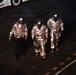 Three individuals wearing reflective silver/aluminized proximity suits and visored helmets are walking across a dark tarmac or pavement surface at night. The suits are consistent with proximity fire suits used by firefighters or hazmat personnel. A vehicle with a visible identification code 'GBQD 000092' is partially visible in the upper left corner.