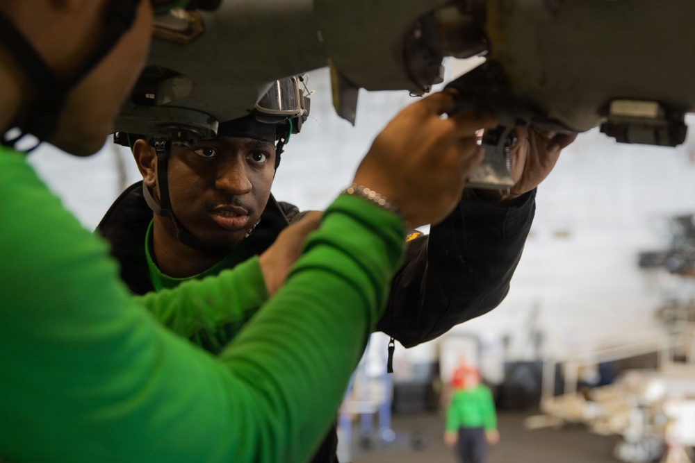 Two individuals wearing green shirts are working on the underside of what appears to be military aircraft equipment in a large hangar bay. The person in focus is wearing a helmet with attached equipment and appears to be closely inspecting or adjusting a component. The setting appears to be a military vessel or air base hangar with additional personnel visible in the background.