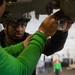 Two individuals wearing green shirts are working on the underside of what appears to be military aircraft equipment in a large hangar bay. The person in focus is wearing a helmet with attached equipment and appears to be closely inspecting or adjusting a component. The setting appears to be a military vessel or air base hangar with additional personnel visible in the background.