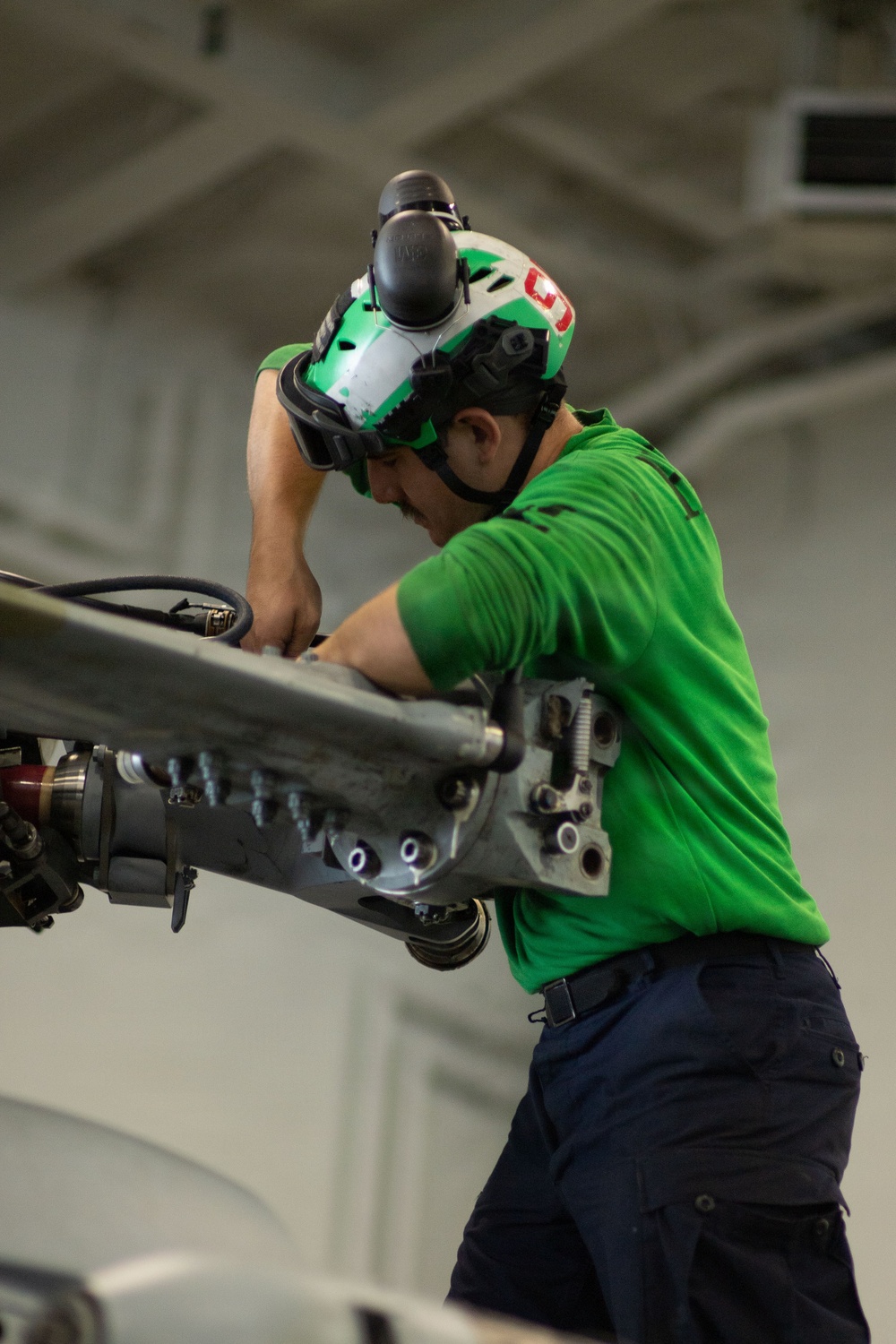 A military or naval aviation technician wearing a green jersey and green/white helmet is performing maintenance work on aircraft equipment in what appears to be a hangar bay. The individual is leaning over and working hands-on with a mechanical component, likely part of an aircraft landing gear, catapult system, or similar aviation hardware. The setting and green jersey are consistent with U.S. Navy carrier aviation maintenance operations.