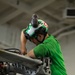 A military or naval aviation technician wearing a green jersey and green/white helmet is performing maintenance work on aircraft equipment in what appears to be a hangar bay. The individual is leaning over and working hands-on with a mechanical component, likely part of an aircraft landing gear, catapult system, or similar aviation hardware. The setting and green jersey are consistent with U.S. Navy carrier aviation maintenance operations.