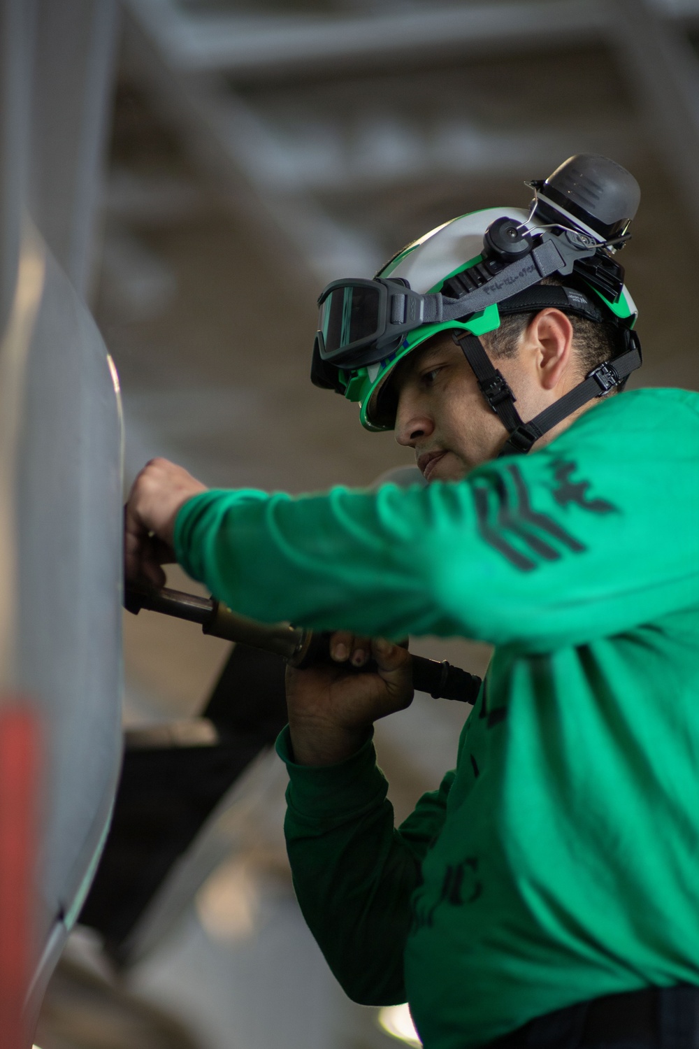 A person wearing a green jersey and a green safety helmet with mounted goggles is performing a technical task, applying force to a tool against a large metallic surface. The individual appears to be working in an industrial or military environment, possibly aboard a naval vessel given the structural elements visible in the background. The green uniform and helmet coloring are consistent with U.S. Navy aviation ordnance or catapult/arresting gear crew color coding.