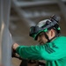 A person wearing a green jersey and a green safety helmet with mounted goggles is performing a technical task, applying force to a tool against a large metallic surface. The individual appears to be working in an industrial or military environment, possibly aboard a naval vessel given the structural elements visible in the background. The green uniform and helmet coloring are consistent with U.S. Navy aviation ordnance or catapult/arresting gear crew color coding.