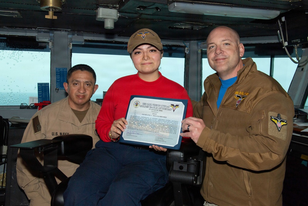 Three individuals are posing for a photo aboard what appears to be a U.S. Navy vessel's bridge or command area. The person in the center is holding a certificate that reads 'Sailor of the Day' dated 19 August 2020, awarded to 'Jackeline C. Noguez-Bruyier' from USS Carl Vinson (CVN-70). Two personnel in military attire flank the award recipient.