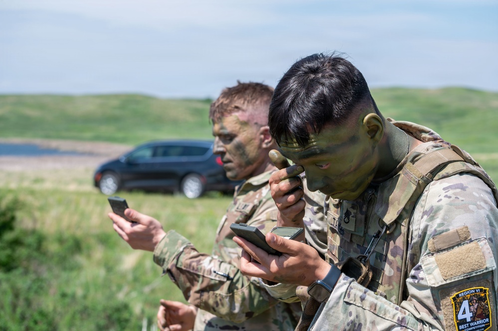 Two military soldiers in camouflage uniforms and face paint are looking at handheld devices, likely smartphones or GPS units, in an outdoor field setting. Both wear OCP (Operational Camouflage Pattern) uniforms and have tactical gear. A patch on the foreground soldier's sleeve reads 'Region VI Best Warrior Competition 4', indicating participation in a military skills competition.
