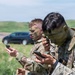 Two military soldiers in camouflage uniforms and face paint are looking at handheld devices, likely smartphones or GPS units, in an outdoor field setting. Both wear OCP (Operational Camouflage Pattern) uniforms and have tactical gear. A patch on the foreground soldier's sleeve reads 'Region VI Best Warrior Competition 4', indicating participation in a military skills competition.