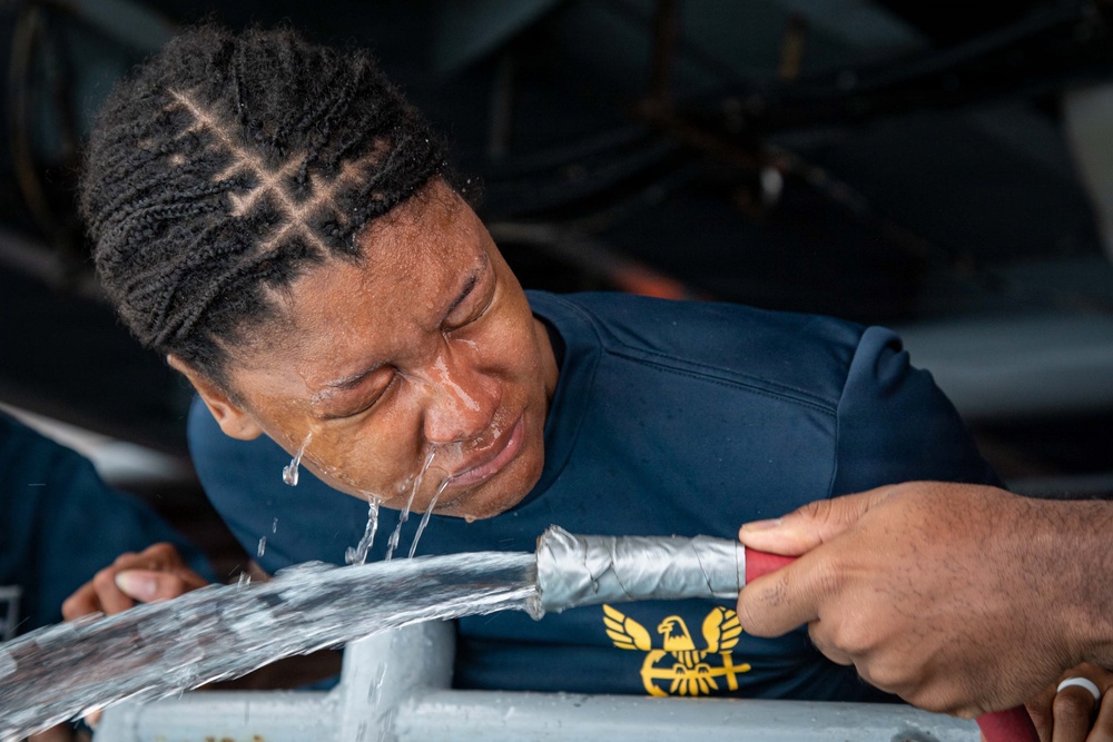 A person wearing a navy blue shirt with a gold eagle/anchor insignia is having their eyes flushed with water from a hose. The water is streaming directly onto their face while their eyes are closed. This appears to be an emergency eye-wash procedure or decontamination protocol, likely in a military or industrial setting.