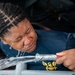 A person wearing a navy blue shirt with a gold eagle/anchor insignia is having their eyes flushed with water from a hose. The water is streaming directly onto their face while their eyes are closed. This appears to be an emergency eye-wash procedure or decontamination protocol, likely in a military or industrial setting.