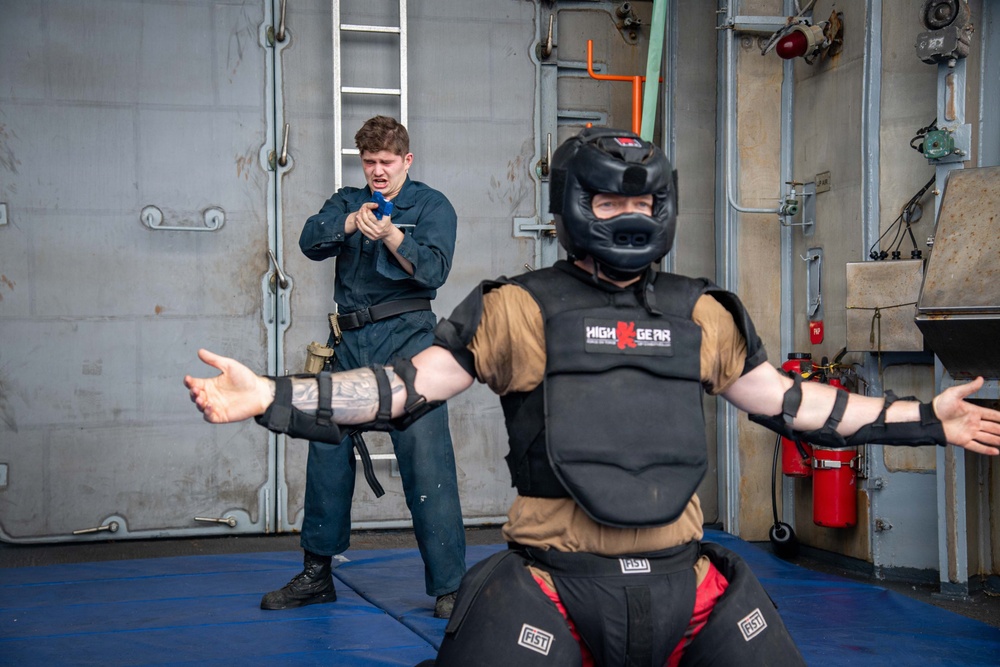 Two individuals are engaged in a combat or self-defense training exercise in what appears to be a military vessel's interior space. One person wears full protective gear including a 'High Gear' brand helmet and 'FIST' brand body armor with arm guards, while the other is dressed in a dark naval coverall uniform. The person in the coverall appears to be reacting to or simulating use of a small blue object, possibly a training weapon or OC spray canister.