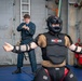 Two individuals are engaged in a combat or self-defense training exercise in what appears to be a military vessel's interior space. One person wears full protective gear including a 'High Gear' brand helmet and 'FIST' brand body armor with arm guards, while the other is dressed in a dark naval coverall uniform. The person in the coverall appears to be reacting to or simulating use of a small blue object, possibly a training weapon or OC spray canister.