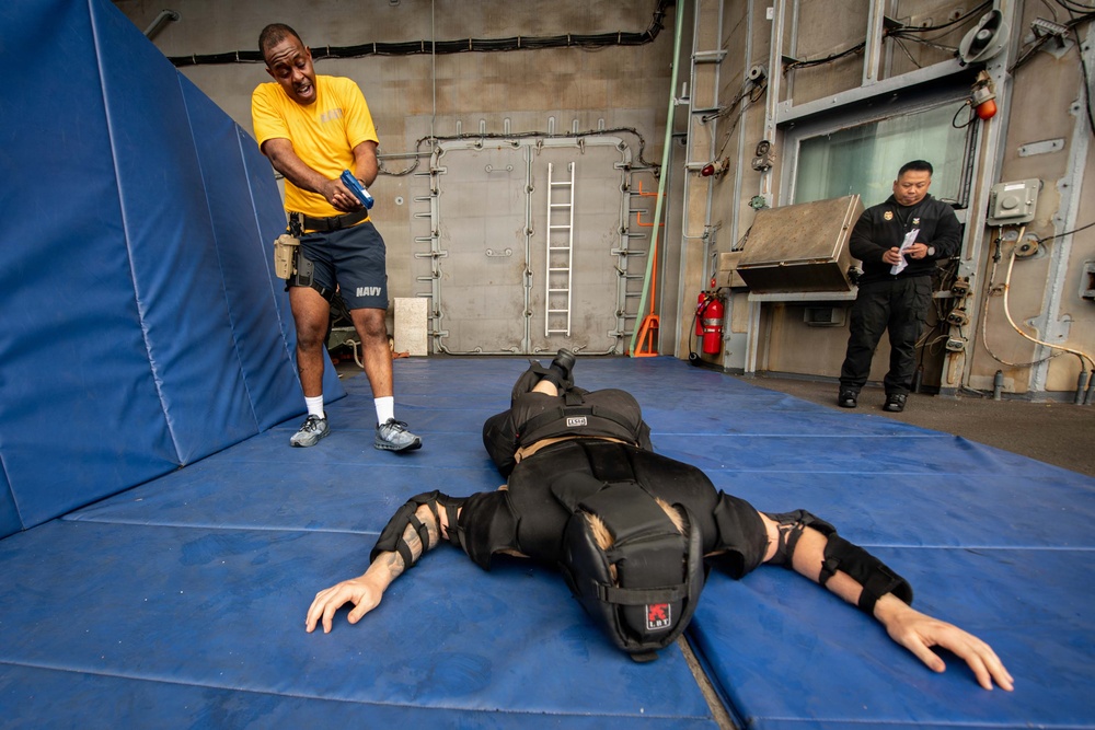 A U.S. Navy training exercise is taking place on blue padded mats inside what appears to be a ship's interior. A sailor in a yellow 'NAVY' shirt is pointing a device (likely a training Taser/OC spray) at a person lying face-down on the mats wearing full protective/padded training gear. A second person in black clothing observes and takes notes in the background.