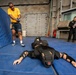 A U.S. Navy training exercise is taking place on blue padded mats inside what appears to be a ship's interior. A sailor in a yellow 'NAVY' shirt is pointing a device (likely a training Taser/OC spray) at a person lying face-down on the mats wearing full protective/padded training gear. A second person in black clothing observes and takes notes in the background.