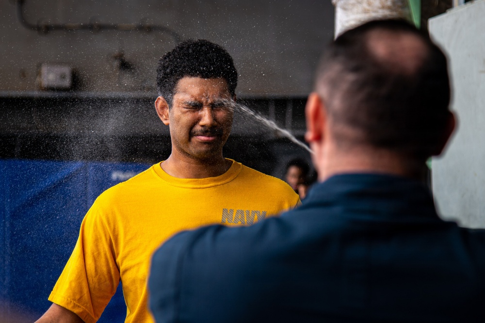 A person wearing a yellow 'NAVY' t-shirt is being sprayed in the face with what appears to be a liquid substance, likely OC (pepper) spray or water during a military/law enforcement training exercise. A second individual in a dark uniform and white cap is administering the spray from the foreground. The subject's eyes are tightly shut and face is contorted in reaction to the spray.