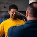A person wearing a yellow 'NAVY' t-shirt is being sprayed in the face with what appears to be a liquid substance, likely OC (pepper) spray or water during a military/law enforcement training exercise. A second individual in a dark uniform and white cap is administering the spray from the foreground. The subject's eyes are tightly shut and face is contorted in reaction to the spray.
