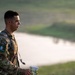 A young male soldier in US Army MultiCam (OCP) camouflage uniform is photographed in profile against a soft, blurred outdoor background featuring a body of water and green fields. He is wearing a tactical backpack and holding what appears to be a map or document, suggesting he is engaged in a land navigation or field exercise. A patch on his left sleeve reads 'Region VI Best Warrior Competition' with the number 8.