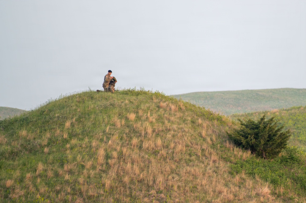 A single individual wearing camouflage clothing stands atop a grassy hill in a rolling landscape, appearing to handle or adjust equipment. The scene is set in an open, hilly terrain with mixed green and golden-brown grasses under an overcast sky. The figure is small relative to the landscape, suggesting the photo was taken from a significant distance.