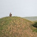 A single individual wearing camouflage clothing stands atop a grassy hill in a rolling landscape, appearing to handle or adjust equipment. The scene is set in an open, hilly terrain with mixed green and golden-brown grasses under an overcast sky. The figure is small relative to the landscape, suggesting the photo was taken from a significant distance.