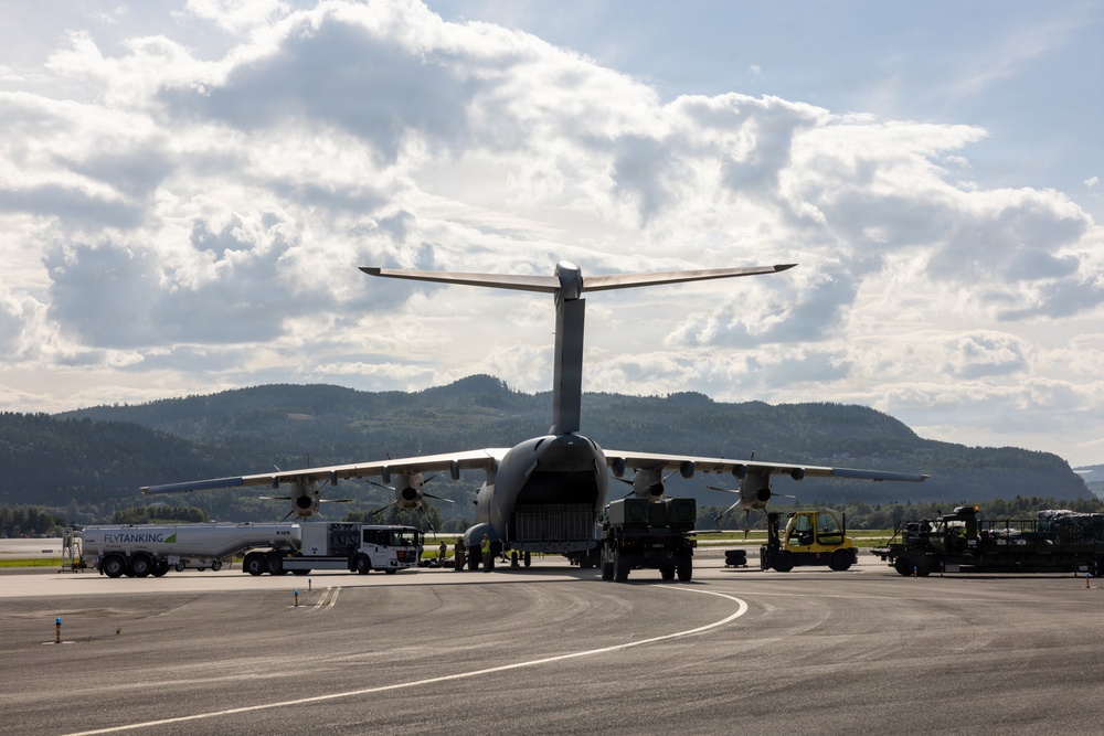 A large military transport aircraft, consistent with an Airbus A400M Atlas, is parked on an airport apron and being serviced with ground support equipment. The aircraft's rear cargo ramp appears open, with loading/unloading operations underway. The setting appears to be a Scandinavian airport, suggested by the forested hills and a fuel truck marked 'FLYTANKING' visible on the left.