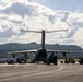 A large military transport aircraft, consistent with an Airbus A400M Atlas, is parked on an airport apron and being serviced with ground support equipment. The aircraft's rear cargo ramp appears open, with loading/unloading operations underway. The setting appears to be a Scandinavian airport, suggested by the forested hills and a fuel truck marked 'FLYTANKING' visible on the left.
