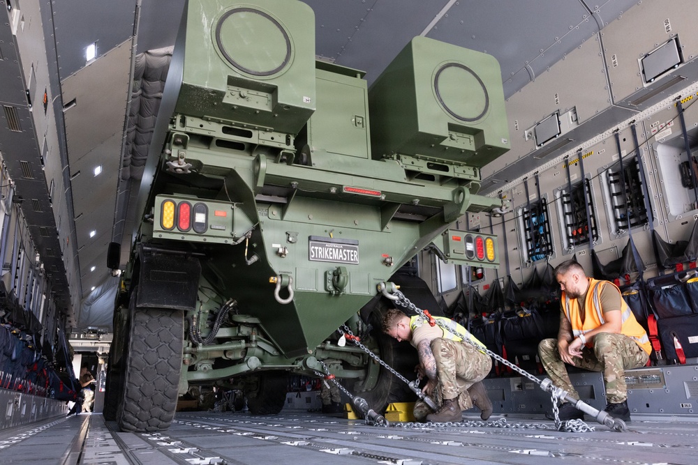 Two military personnel in combat uniforms and high-visibility orange vests are securing a military vehicle labeled 'STRIKEMASTER' inside the cargo hold of a large military transport aircraft. The vehicle appears to be a wheeled armored or weapons platform being chained down to the aircraft floor using heavy-duty tie-down chains. The interior of the aircraft shows standard military cargo configuration with tie-down rails, equipment storage panels, and overhead lighting.