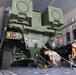 Two military personnel in combat uniforms and high-visibility orange vests are securing a military vehicle labeled 'STRIKEMASTER' inside the cargo hold of a large military transport aircraft. The vehicle appears to be a wheeled armored or weapons platform being chained down to the aircraft floor using heavy-duty tie-down chains. The interior of the aircraft shows standard military cargo configuration with tie-down rails, equipment storage panels, and overhead lighting.