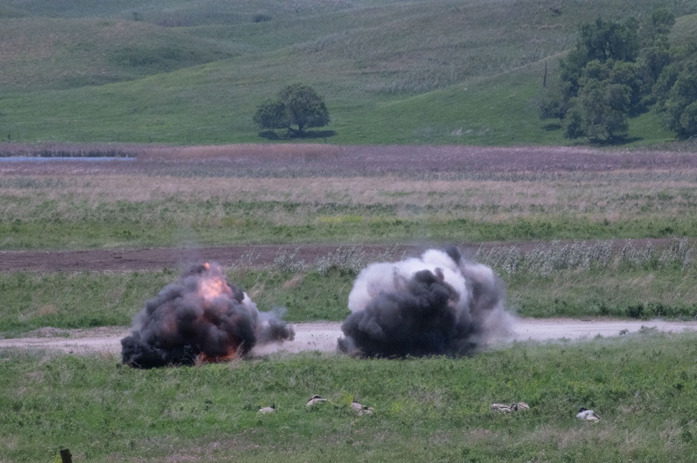The image shows two simultaneous explosions occurring on an open grassy field or training range. The left explosion displays visible orange flames with dark black smoke, while the right explosion produces a larger gray-white dust and smoke cloud. The setting appears to be a military training exercise or controlled demolition on a rural landscape.
