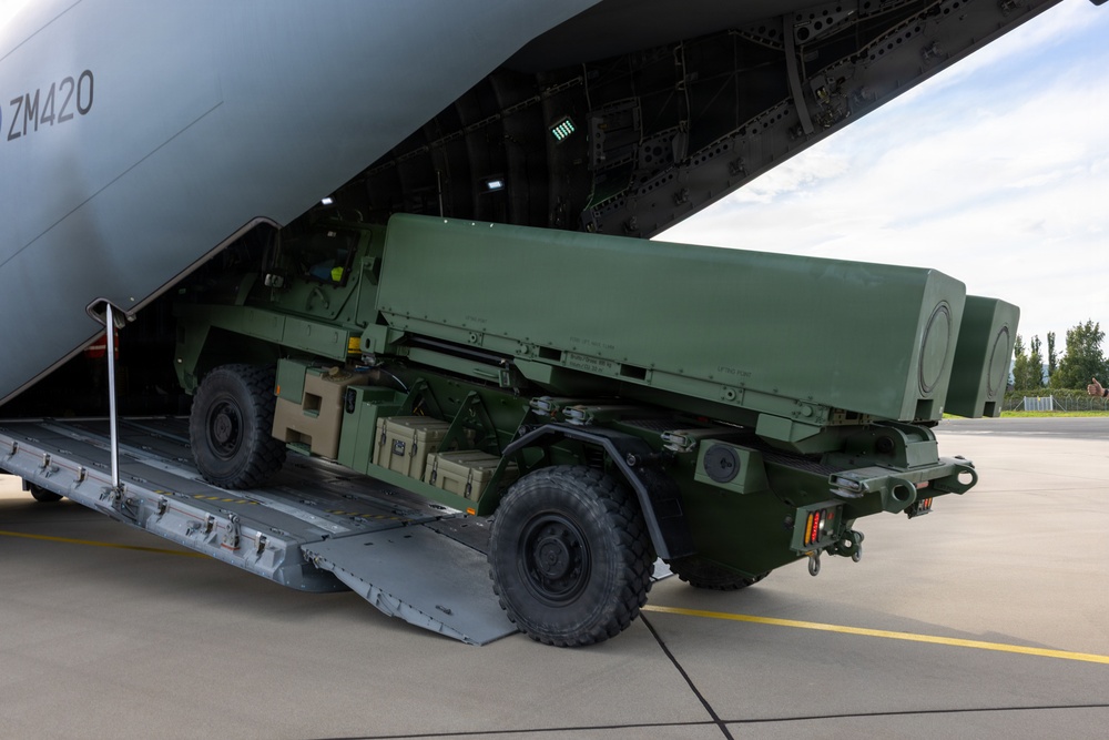 A military wheeled vehicle, consistent with a HIMARS (High Mobility Artillery Rocket System) launcher, is being loaded or unloaded via a rear cargo ramp of a large military transport aircraft marked 'ZM420'. The aircraft appears to be a Royal Air Force Atlas A400M based on the tail number format and fuselage color. The operation is taking place on a paved airfield apron under partly cloudy skies.