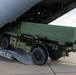 A military wheeled vehicle, consistent with a HIMARS (High Mobility Artillery Rocket System) launcher, is being loaded or unloaded via a rear cargo ramp of a large military transport aircraft marked 'ZM420'. The aircraft appears to be a Royal Air Force Atlas A400M based on the tail number format and fuselage color. The operation is taking place on a paved airfield apron under partly cloudy skies.