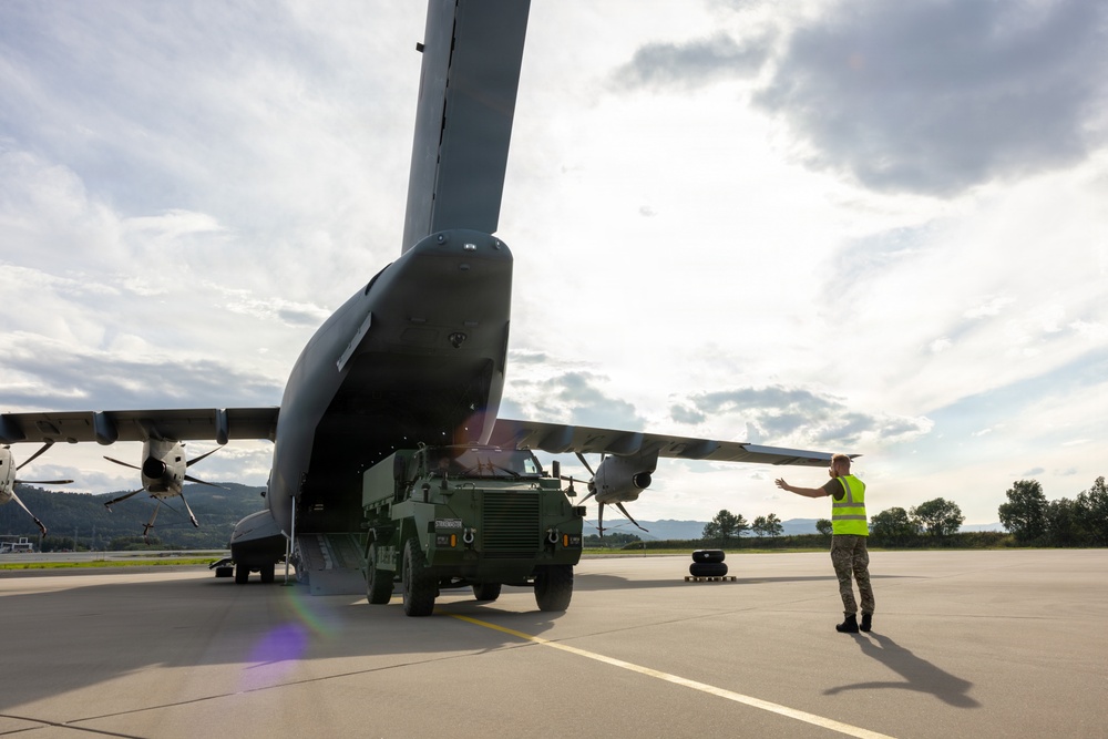 A military ground crew member in a high-visibility vest is marshalling a military tactical vehicle as it is loaded or unloaded from the rear ramp of a large military transport aircraft on a tarmac. The scene appears to take place at a military airfield surrounded by hills and trees. The image captures a standard military airlift logistics operation in progress.