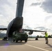 A military ground crew member in a high-visibility vest is marshalling a military tactical vehicle as it is loaded or unloaded from the rear ramp of a large military transport aircraft on a tarmac. The scene appears to take place at a military airfield surrounded by hills and trees. The image captures a standard military airlift logistics operation in progress.
