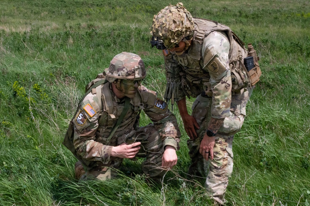 Two U.S. Army soldiers in Operational Camouflage Pattern (OCP) uniforms are crouched in a grassy field, examining something on the ground. Both wear tactical gear including plate carriers, helmets, and unit patches including American flag patches. They appear to be conducting a field training exercise or reconnaissance activity.