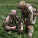 Two U.S. Army soldiers in Operational Camouflage Pattern (OCP) uniforms are crouched in a grassy field, examining something on the ground. Both wear tactical gear including plate carriers, helmets, and unit patches including American flag patches. They appear to be conducting a field training exercise or reconnaissance activity.