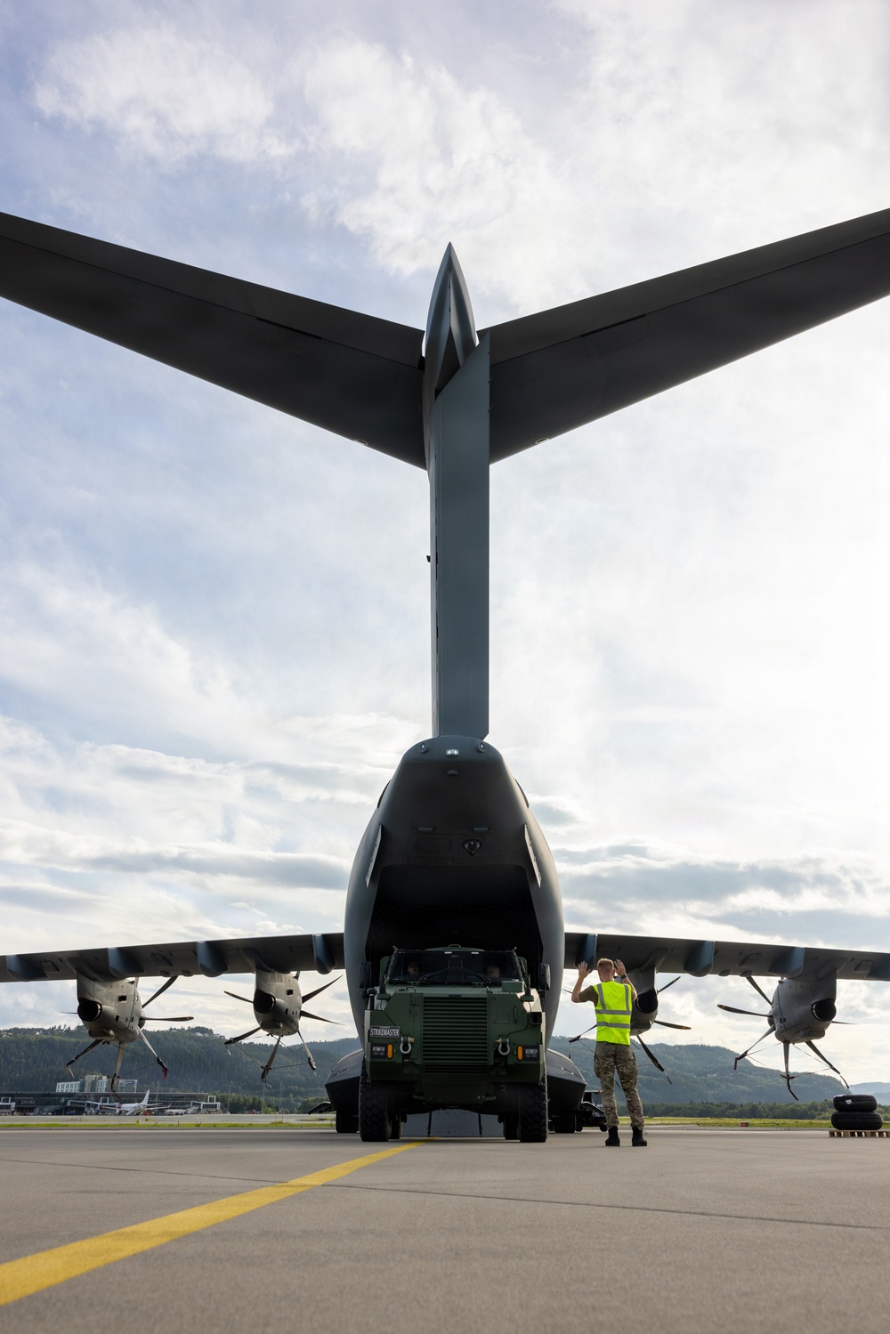 A rear-view photograph of a large military transport aircraft with its cargo ramp open, as a military ground vehicle is being marshaled out of or into the aircraft. A ground crew member wearing a high-visibility yellow vest is directing the vehicle using hand signals. The image is taken from directly behind the aircraft on a paved tarmac, with forested hills and partial airport infrastructure visible in the background.