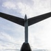 A rear-view photograph of a large military transport aircraft with its cargo ramp open, as a military ground vehicle is being marshaled out of or into the aircraft. A ground crew member wearing a high-visibility yellow vest is directing the vehicle using hand signals. The image is taken from directly behind the aircraft on a paved tarmac, with forested hills and partial airport infrastructure visible in the background.