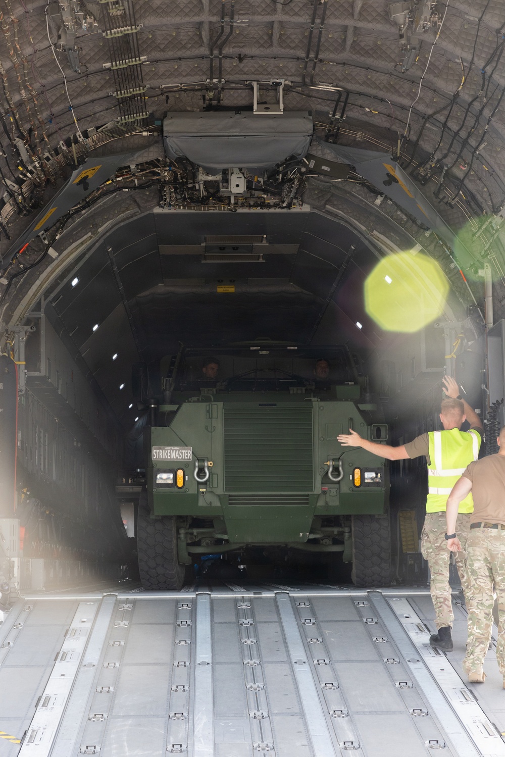 A green military armored vehicle labeled 'STRIKEMASTER' is being loaded or unloaded from a large military transport aircraft, likely a C-17 Globemaster III or similar heavy-lift aircraft. Ground crew personnel in high-visibility vests and military camouflage uniforms are guiding the vehicle through the aircraft's rear cargo ramp. The scene appears to be an Australian military logistics or training operation based on the vehicle markings.