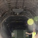 A green military armored vehicle labeled 'STRIKEMASTER' is being loaded or unloaded from a large military transport aircraft, likely a C-17 Globemaster III or similar heavy-lift aircraft. Ground crew personnel in high-visibility vests and military camouflage uniforms are guiding the vehicle through the aircraft's rear cargo ramp. The scene appears to be an Australian military logistics or training operation based on the vehicle markings.