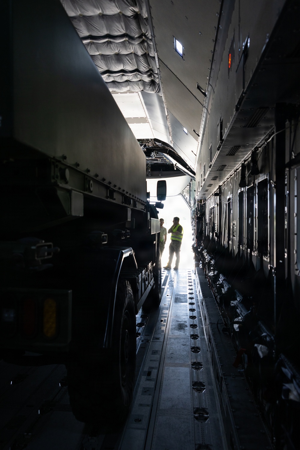 The image shows the interior cargo hold of a large military or heavy-lift transport aircraft, with a large wheeled vehicle loaded inside. Two personnel are visible near the rear ramp opening, silhouetted against bright exterior light. The scene appears to depict a cargo loading or inspection operation.
