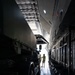 The image shows the interior cargo hold of a large military or heavy-lift transport aircraft, with a large wheeled vehicle loaded inside. Two personnel are visible near the rear ramp opening, silhouetted against bright exterior light. The scene appears to depict a cargo loading or inspection operation.