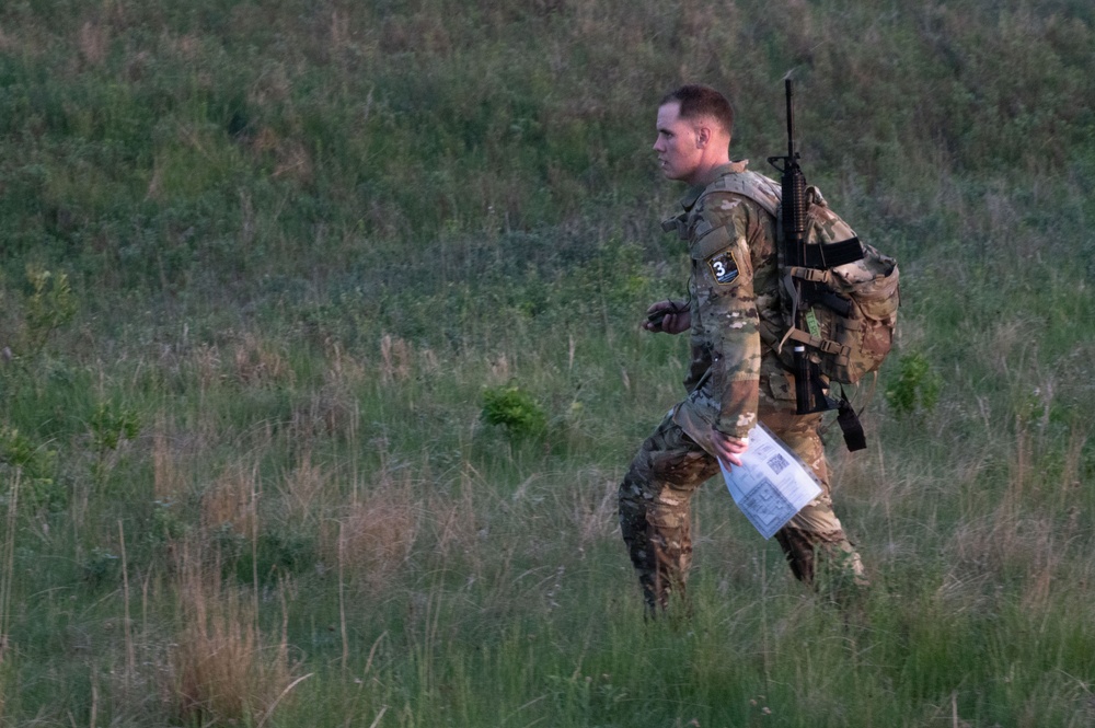 A soldier in full camouflage uniform is moving through an open grassy field, carrying a rifle on their back and holding what appears to be a map or document. The individual wears a numbered bib or identifier marked '3', suggesting participation in a military competition, exercise, or land navigation event. The setting appears to be a training area during early morning or evening light.