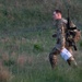 A soldier in full camouflage uniform is moving through an open grassy field, carrying a rifle on their back and holding what appears to be a map or document. The individual wears a numbered bib or identifier marked '3', suggesting participation in a military competition, exercise, or land navigation event. The setting appears to be a training area during early morning or evening light.
