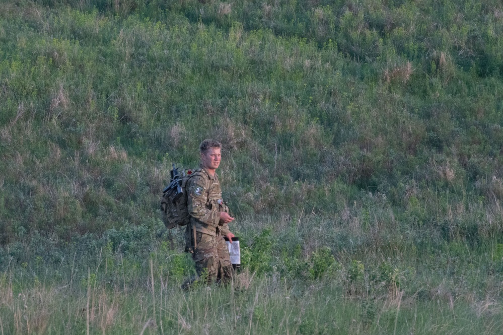 A young male soldier in camouflage uniform stands in an open grassy field with a hillside covered in shrubs and vegetation behind him. He is carrying a tactical backpack and holding what appears to be a map or document, suggesting he is engaged in a land navigation exercise. The image was taken from a distance with a telephoto lens.