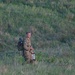A young male soldier in camouflage uniform stands in an open grassy field with a hillside covered in shrubs and vegetation behind him. He is carrying a tactical backpack and holding what appears to be a map or document, suggesting he is engaged in a land navigation exercise. The image was taken from a distance with a telephoto lens.